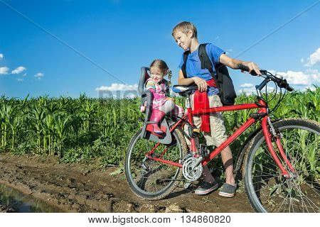 Sibling teenage boy is cycling with his little sibling sister on baby bike seat on farm corn field dirt road