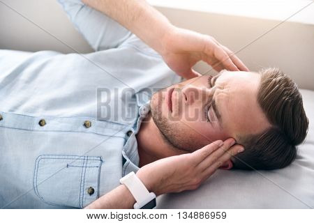 Severe disease. Cheerless sick man lying on the sofa and holding his hands on the temples while having a headache
