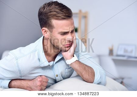 You should visit your doctor. Cheerless sick man sitting on the couch and feeling pain while holding hand on the face