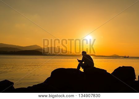 Young man sitting on the stone play smartphone at sea sunset background
