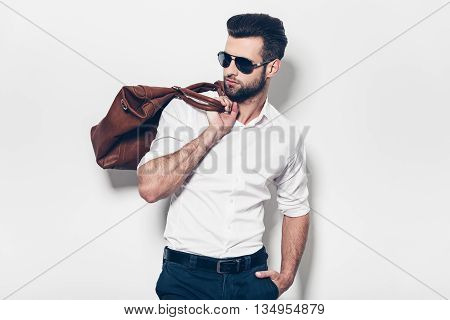 Ready to go. Handsome young man in white shirt carrying leather bag on shoulder and looking away while standing against white background