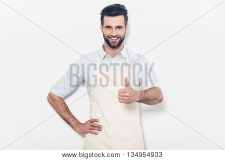 Good job! Confident young handsome man in apron keeping arms crossed and smiling while standing against white background