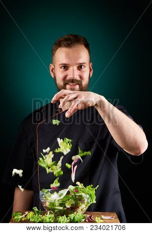 Portrait Of A Handsome Mischevious Male Chef Cook In Black Uniform Making Delicious Vegetable Salad 