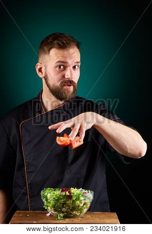 Portrait Of A Handsome Mischevious Male Chef Cook In Black Uniform Making Delicious Vegetable Salad 