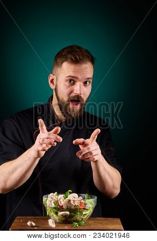 Portrait Of A Handsome Mischevious Male Chef Cook In Black Uniform Making Delicious Vegetable Salad 