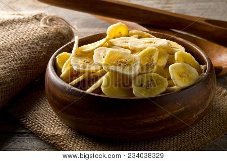 Heap Of Dried Banana Chips Snack In Wooden Bowl On Burlap On Dark Wooden Table