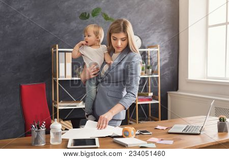 Happy Mother Working At Office With Her Baby. Young Woman Reading Documents While Spending Time With