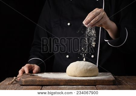 Chef Hands Pouring Flour Powder On Raw Dough Using Sieve On A Black Background, Cooking Process.