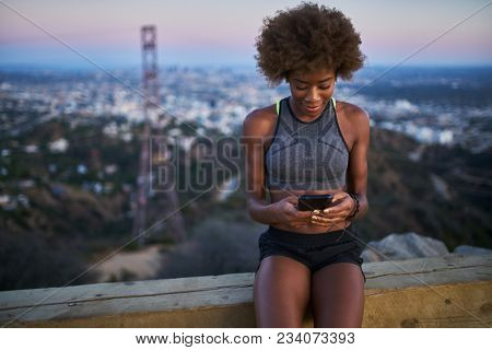 athletic african american woman using smartphone at runyon canyon while resting on bench at dusk
