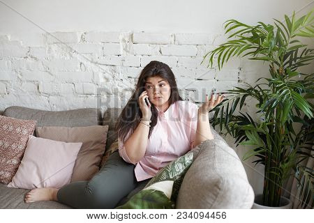 Astonished Indignant Plus Size Young Brunette Female Sitting In Living Room, Gesturing Emotionally W