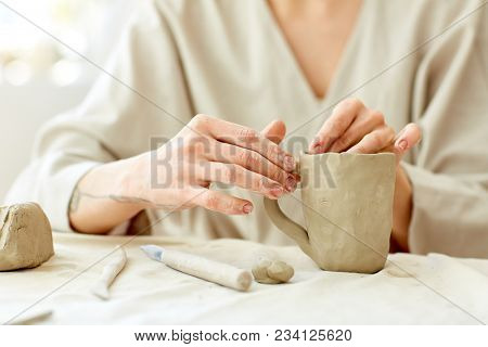 Hands of artisan making clay or ceramic mug by workplace with necessary handtools near by