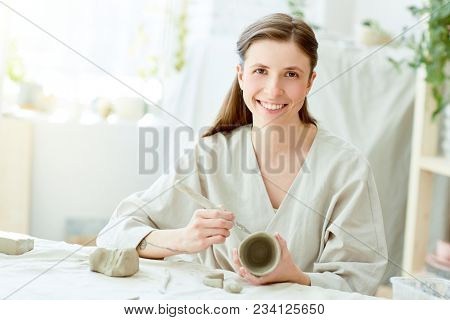 Smiling girl holding self-made clay mug and handtool for cutting ornaments and looking at camera by her workplace