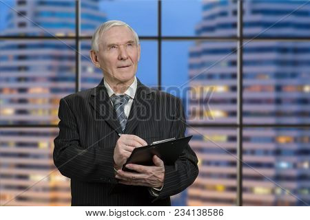 Senior Man In Black Suit Writing On Clipboard Paper. Looking Up And Writing Down. Inside Skyscrapper