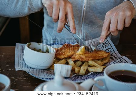 Man Sitting At The Wooden Table, Eating Fish And Chips, Traditional English Dish, Tea And Sauces Nea
