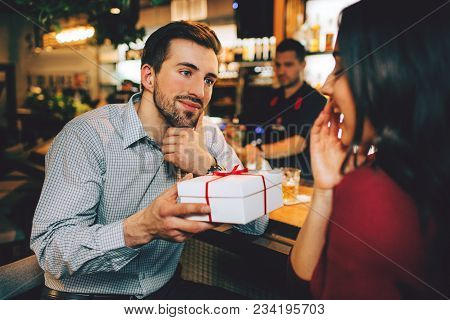 Boyfriend And Girlfriend Sitting Together In Restaurant. He Has A Present For Her. She Didn't Expect