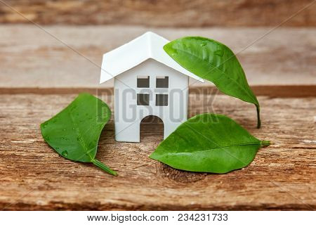 Miniature White Toy Model House With Green Leaves On Wooden Backgdrop. Eco Village, Abstract Environ