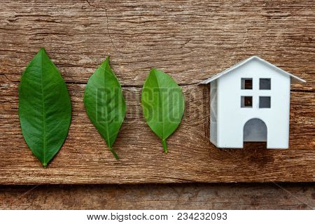 Miniature White Toy Model House With Green Leaves On Wooden Backgdrop. Eco Village, Abstract Environ