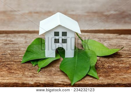 Miniature White Toy Model House With Green Leaves On Wooden Backgdrop. Eco Village, Abstract Environ