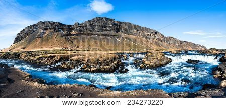 Fossalar River Rapids In Rural Iceland With Mountains, A Farmhouse, Grass, And Blue Sky
