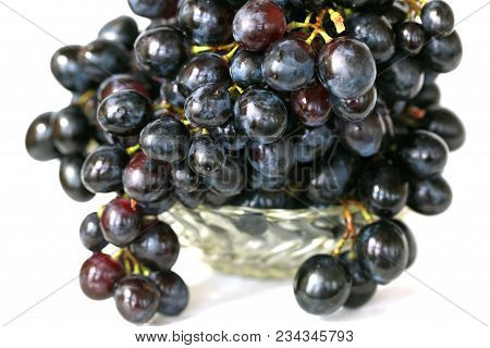A Bunch Of Drooping Black Grapes In A Small Glass Vase.