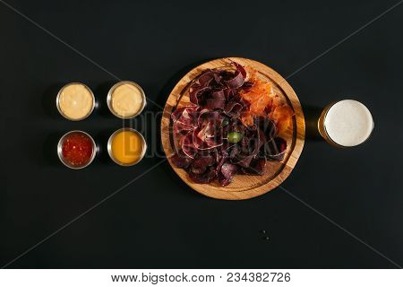 Top View Of Delicious Sliced Assorted Meat On Wooden Board With Glass Of Beer And Sauces On Black