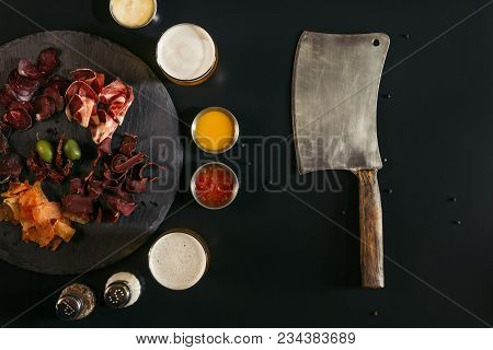 Top View Of Meat Knife And Gourmet Sliced Assorted Meat On Slate Board With Sauces And Spices On Bla