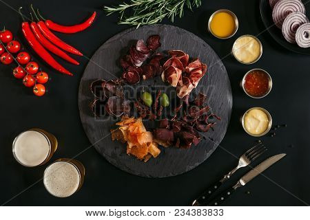Gourmet Assorted Meat On Slate Board, Various Sauces, Vegetables And Glasses Of Beer On Black