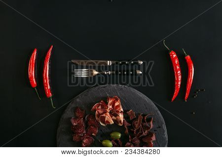 Top View Of Gourmet Assorted Meat On Slate Board, Fork With Knife And Chili Peppers On Black