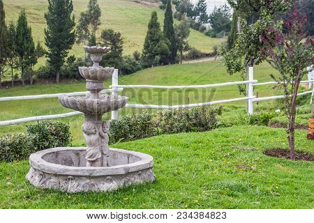 View Of Fountain In The Garden With White Fence, Pastures, And Hills