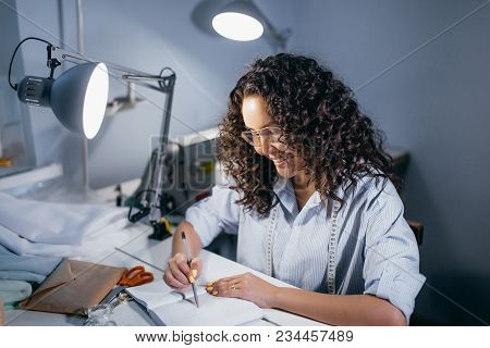 Side View Photo Of Female Dressmaker Bending Over The Table With Diary, Envelope, Scissors And Fabri
