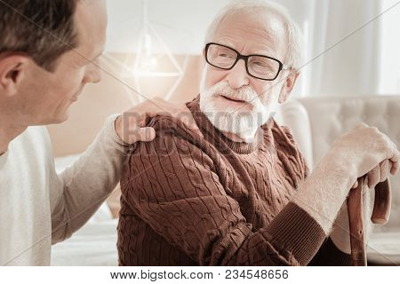 Fathers Care. Bespectacled Attentive Aged Man Sitting On The Bed Supporting On The Cane And Listenin