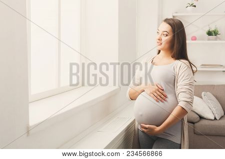 Pregnant Woman Caressing Her Belly At Home. Young Expectant Lady Feeling Her Baby Push, Standing At 