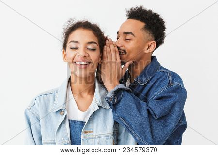 Smiling african man talking something in ear of his pleased girlfriend which listening with closed eyes over grey background