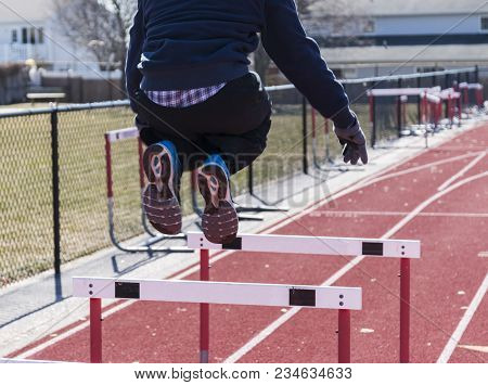 A High School Male Athlette Is Jumping Over Hurdles On A Cold Afternoon During Track And Field Pract