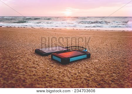 Powerbank In The Sand On Background Of The Rising Sun In The Cloudy Sky And Blue Sea. Alternative En