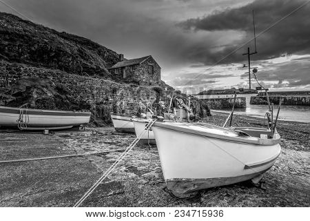 White Boats In Stormy Harbour With Little House On The Hill At Picturesque Mullion Cove, Cornwall, E