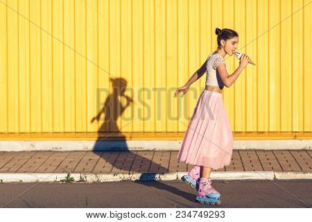 Young Girl On Summer Evening Roller-skating And Eating Ice Cream On Yellow Wall Background. Concept 