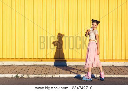 Young Girl On Summer Day Goes Rollerblading And Eats An Ice Cream On The Background Of Yellow Wall. 