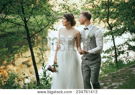 The Bride And Groom Walk On A Background Of Lake. Romantic Wedding Photo.