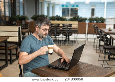 Freelance And Working Online Concept. Man Working With A Black Laptop, While Drinking Latte In A Cof