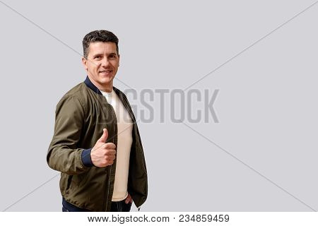 Attractive Smiling Middle-aged Man In A Green  Jacket Stands On A Gray Background And Makes A Gestur