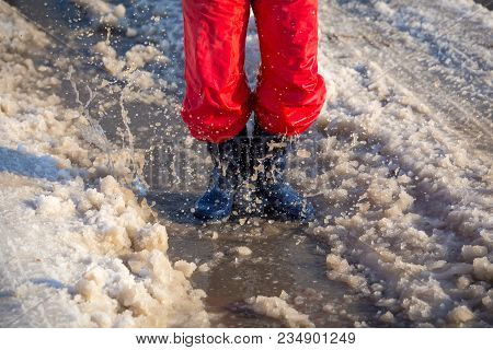 Kid In Blue Rainboots Jumping In The Ice Puddle With Melting Snow At Sunny Spring Day, Outdoors
