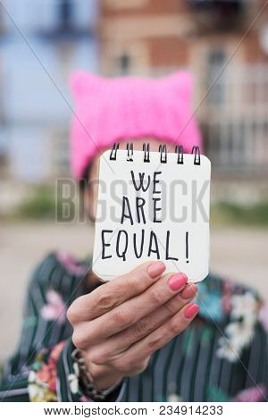 closeup of a young woman outdoors wearing a pink pussycat hat showing a notepad in front of her face with the text we are equal written in it