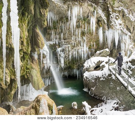 Hiker Man Standing On Bridge Enjoying Icicles And Waterfall On Winter Trekking Hike. Hinanger Waterf