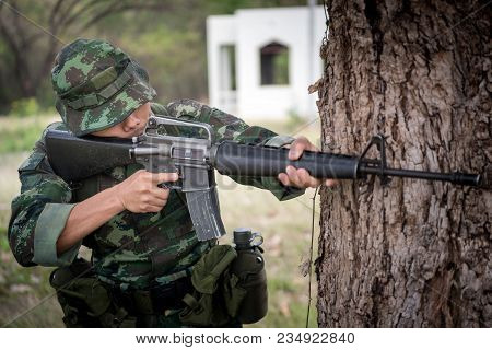 Special Forces Soldier Or Private Millitary Holding Gun Aiming Behind A Tree At Field Area.