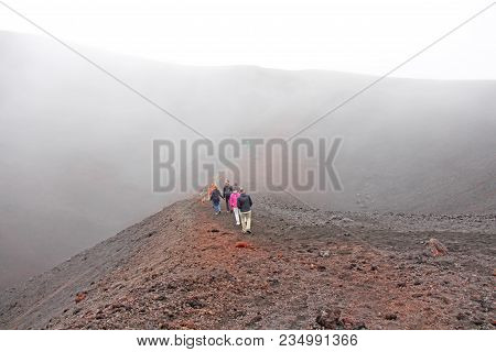 People Walk Along The Crater Of Mount Etna. Black Volcanic Earth And Thick Fog On Mount Etna. Place 