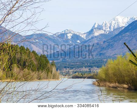 A Serene View On Snowy Mountain From A River Side. The River Is Overgrown With Trees On Both Sides. 