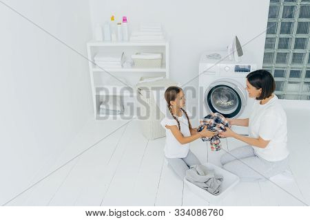 Little Girl Helper And Her Mother Pose In Laundry Room Near Washing Machine, Sit On White Floor, Hol