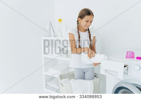 Small Adorable Busy Girl Stands In Laundry Basket, Pours Detergent In Washing Machine Compartment, H