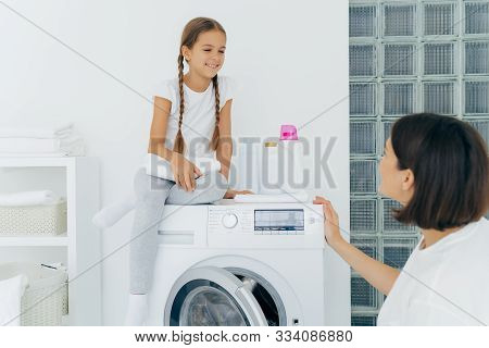 Adorable Small Girl With Pigtails, Poses On Top Of Washer, Holds White Soft Towel, Looks Gladfully A